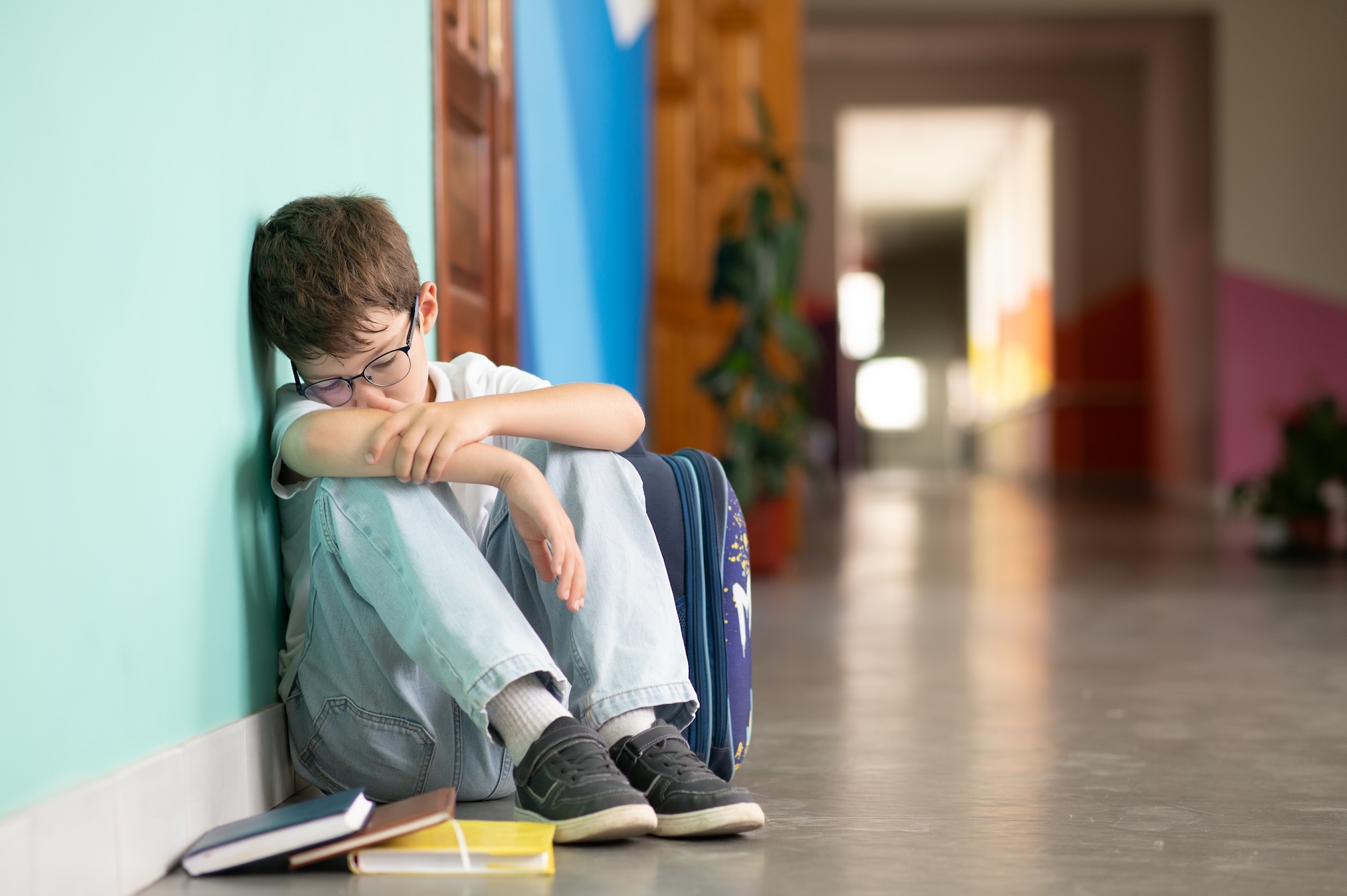 Little boy sitting alone on floor after suffering an act of bullying. Sad young schoolboy sitting on corridor with hands on knees and head between his legs