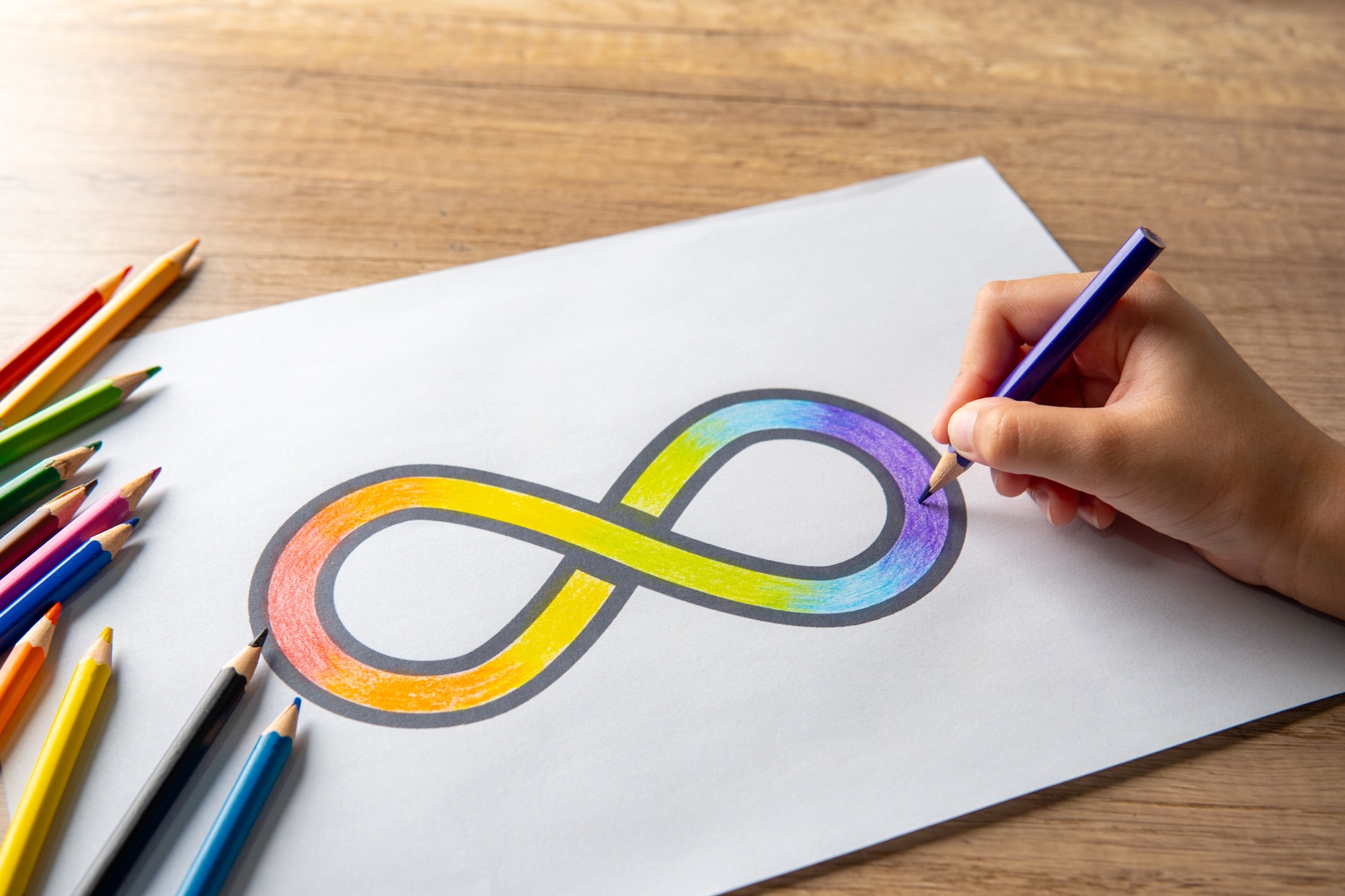 A hand coloring a rainbow infinity symbol on a white sheet of paper with colored pencils scattered around. Autism awareness concept.